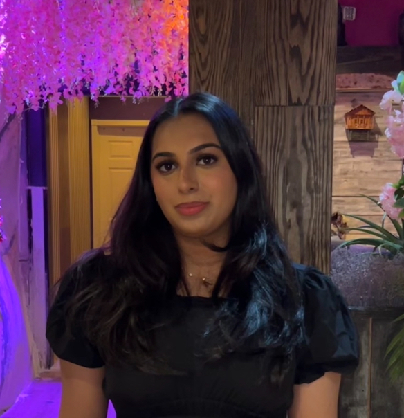 Person with long dark hair wearing a black top, standing in front of a colorful background with purple foliage and a multi-colored doorway.