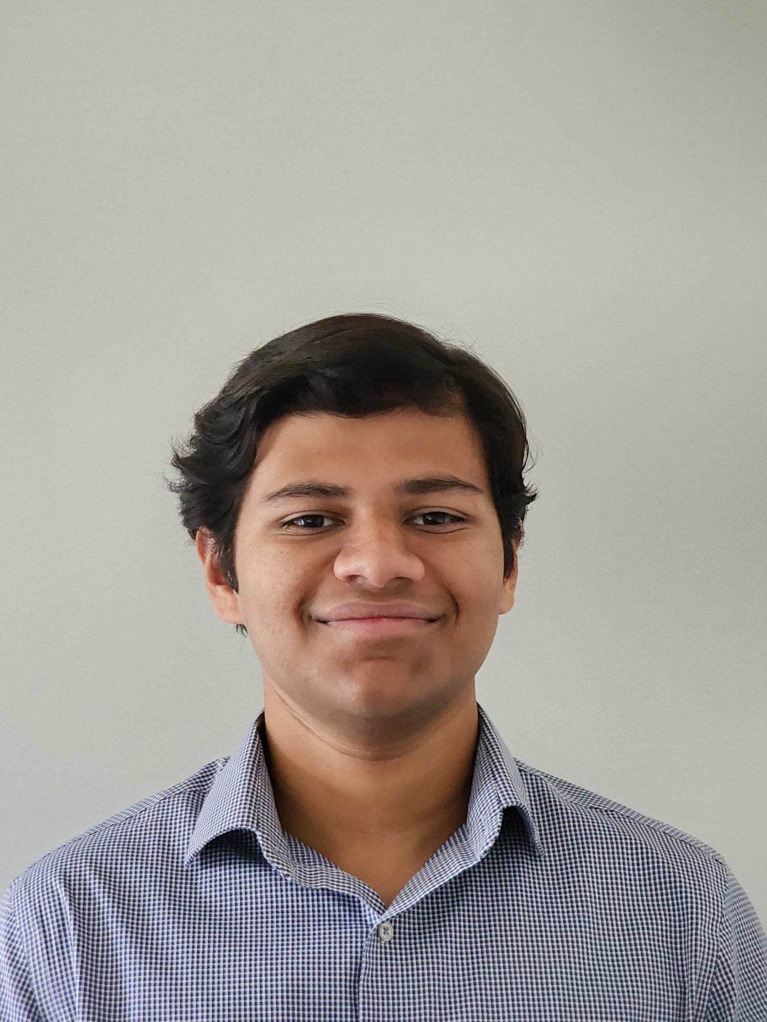 Portrait of a smiling person with short, wavy hair, wearing a blue checked shirt, standing against a plain light gray background.