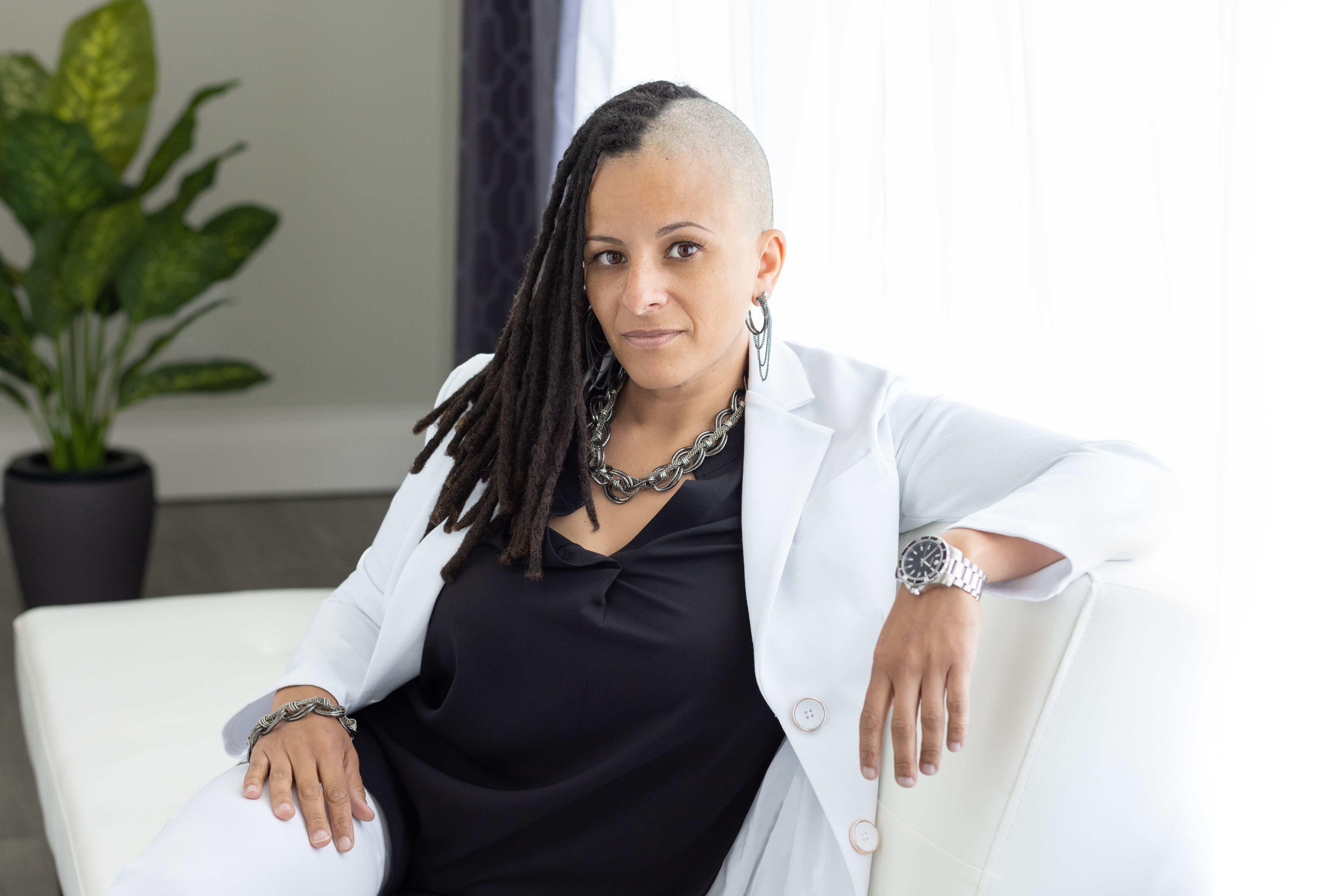 Person with a shaved head and braids wearing a white blazer and black top, sitting confidently on a white sofa in a well-lit room with a potted plant and curtains in the background.