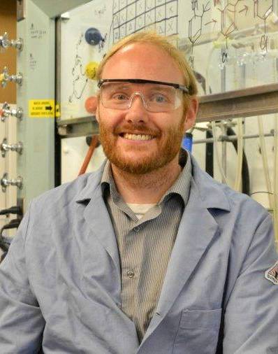 A person wearing safety glasses and a lab coat smiling in front of a wall filled with chemical formulas and laboratory apparatus.