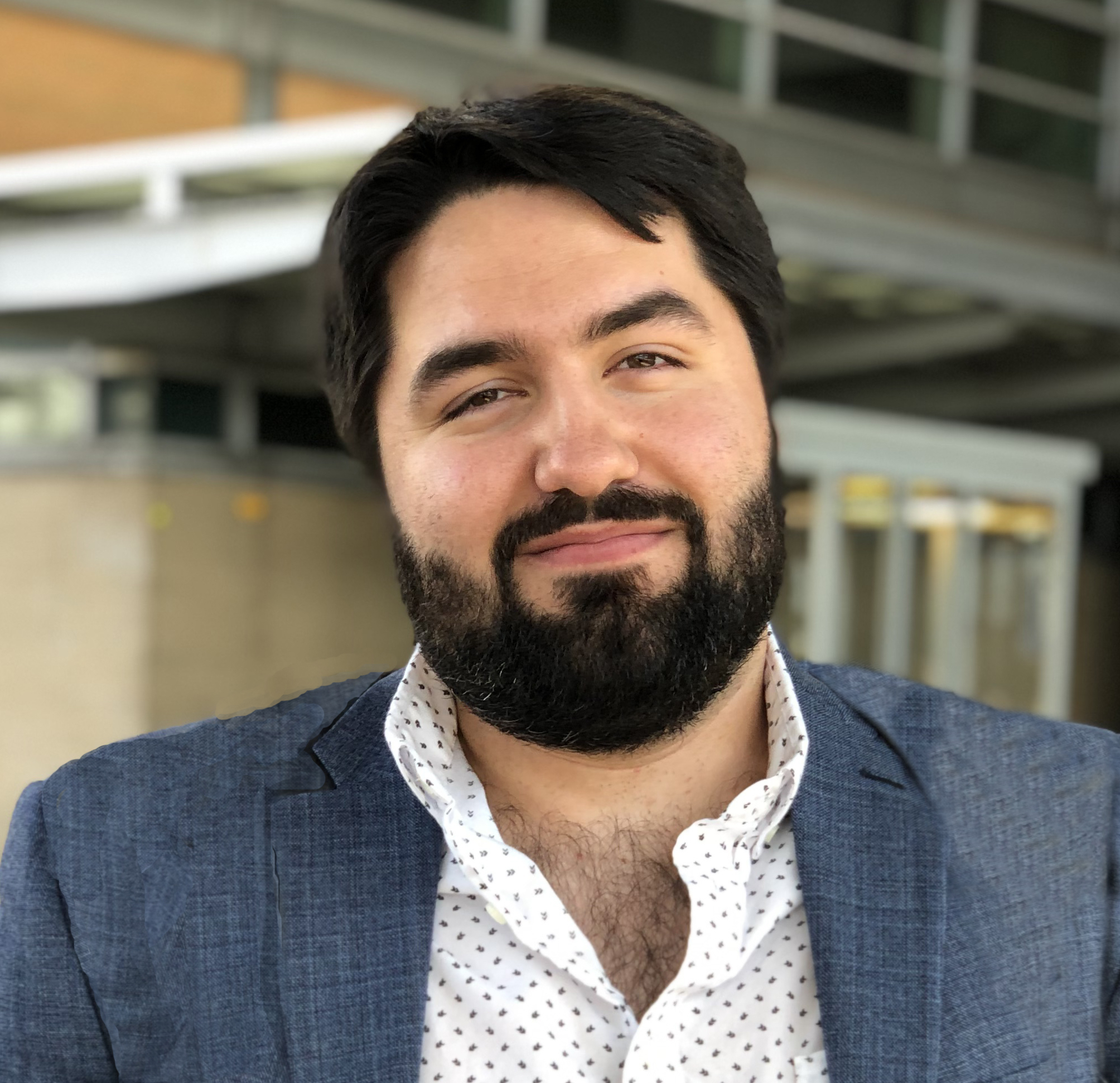 A person with a beard, wearing a blazer and a polka-dot shirt, smiling in front of a building with visible stairs and metal railings in the background.