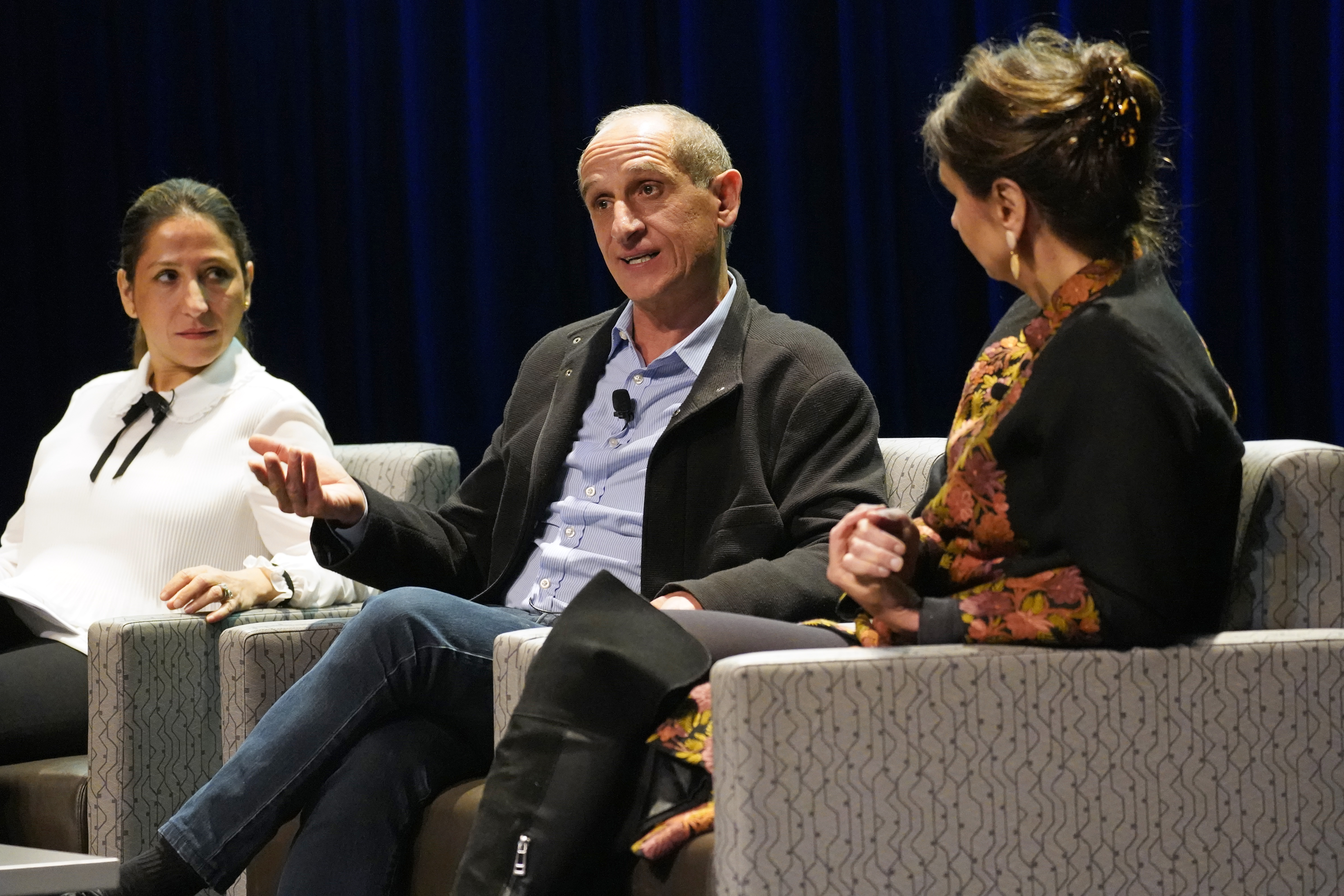 a man speaks from a seat on stage during a panel discussion. he is flanked by two seated women.